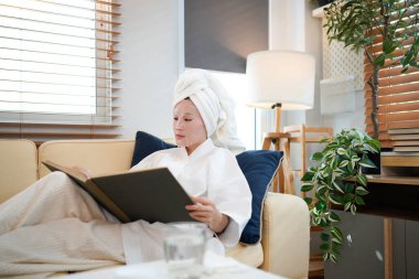 Beyaz bornozlu kadın, kanepede havluya sarılı saçlar, çarşaf maskesi ve rahat bir evde rahatlayan bir kitap. Güzellik ve cilt bakımı estetiği.