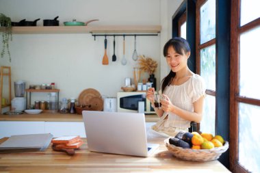 Mutlu serbest çalışan kadın evde çalışıyor, kahve içiyor ve bilgisayarını kullanıyor. Modern mutfakta yemek yerken aynı zamanda sandviç ve meyvelerle kahvaltı yapıyor..