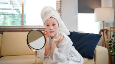 Beyaz bornozlu Asyalı kadın makyaj aynası ve doğal cilt bakımı ile maske takıyor. Güzel bir kadın kendini güzellik tedavileriyle şımartıyor..