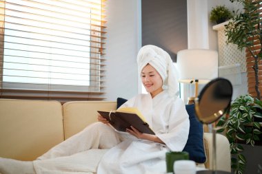 Beyaz bornozlu kadın, kanepede havluya sarılı saçlar ve rahat bir evde kitap dinleniyor. Güzellik ve cilt bakımı estetiği..