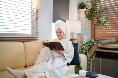 Beyaz bornozlu kadın, kanepede havluya sarılı saçlar ve rahat bir evde kitap dinleniyor. Güzellik ve cilt bakımı estetiği..