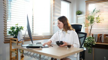 Ofis masasında oturan kadın masaüstü bilgisayarıyla kahvaltı yapıyor. Gülümseyen rahat genç bayan melez modern evdeki bilgisayarda çalışıyor..