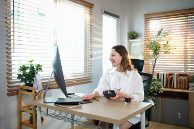 Ofis masasında oturan kadın masaüstü bilgisayarıyla kahvaltı yapıyor. Gülümseyen rahat genç bayan melez modern evdeki bilgisayarda çalışıyor..