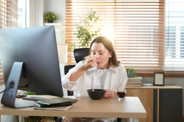 Ofis masasında oturan kadın masaüstü bilgisayarıyla kahvaltı yapıyor. Gülümseyen rahat genç bayan melez modern evdeki bilgisayarda çalışıyor..
