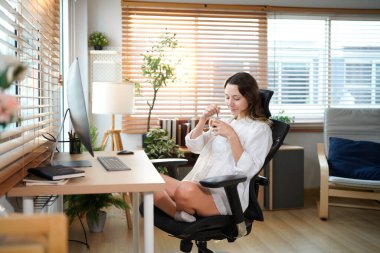 Beyaz bir kadın ofis masasında oturmuş masaüstü bilgisayarıyla kahve içiyor. Gülümseyen rahat genç bayan melez modern evdeki bilgisayarda çalışıyor..