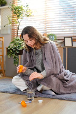 Elinde battaniyeyle yerde oturan genç bir kadın hasta ve grip yüzünden öksürüyor. Evinin oturma odasındaki belirtileri gidermek için ilaç alıyor..