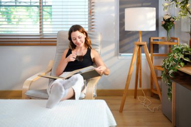 Genç kadın sıcak içecekten hoşlanıyor, kitap okuyor ve pencere ışığında rahat bir şekilde oturuyor..