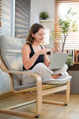 Kahve fincanı ve dizüstü bilgisayarı olan mutlu genç bir kadın evdeki oturma odasında oturuyor..