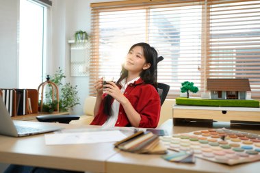 Genç mimar evde rahat bir atmosferde çalışıyor. Gülümseyen Asyalı kadın mimar masada sıcak kahve içiyor, ev ofisinin planlarıyla, bir fincan kahvenin tadını çıkarıyor..