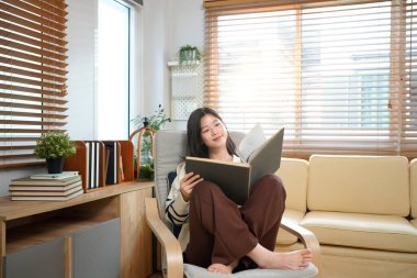 Güzel Asyalı kadın, rahat oturma odasında rahat bir koltukta, bir bitki ve kitaplıkla kitap okuyor, güneşli bir pencere izliyor..