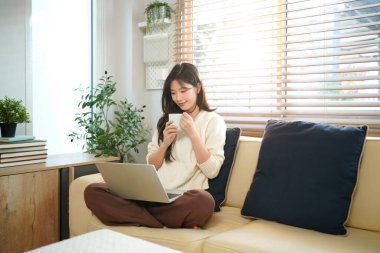 Oturma odasındaki kanepede oturmuş, elinde kupayla dizüstü bilgisayarını kullanan bir kadın gülümsüyor..