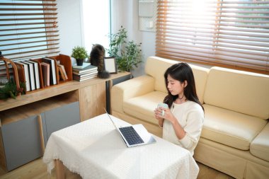 Oturma odasında oturmuş, elinde kupayla dizüstü bilgisayar kullanan bir kadın gülümsüyor..