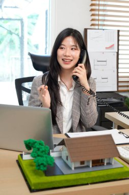 Emlakçı, modern bir ofiste dizüstü bilgisayar ve telefon üzerinde çalışıyor. Masasında da ev modeli var. Emlak projesi planlayan profesyonel bir iş kadını. Konut piyasası.