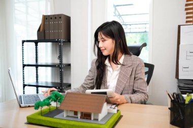 Ev modelli iş ipoteği emlakçısı, masada oturuyor, emlakçı hesap makinesi ve belgeler, ipotek ve mülk üzerinde çalışıyor..