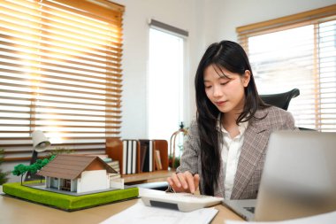 Ev modelli iş ipoteği emlakçısı, masada oturuyor, emlakçı hesap makinesi ve belgeler, ipotek ve mülk üzerinde çalışıyor..