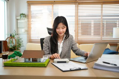 Ev modelli iş ipoteği emlakçısı, masada oturuyor, emlakçı hesap makinesi ve belgeler, ipotek ve mülk üzerinde çalışıyor..