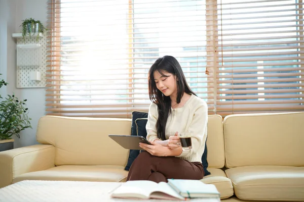 Oturma odasındaki bir kanepede otururken gülümseyen bir kadın elinde bardak ve tablet bir aletle..