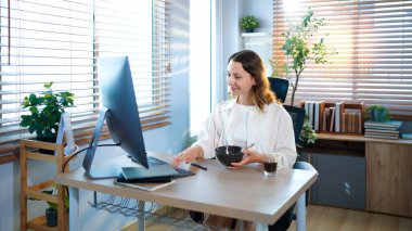 Ofis masasında oturan kadın masaüstü bilgisayarıyla kahvaltı yapıyor. Gülümseyen rahat genç bayan melez modern evdeki bilgisayarda çalışıyor..