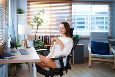 Beyaz bir kadın ofis masasında oturmuş masaüstü bilgisayarıyla kahve içiyor. Gülümseyen rahat genç bayan melez modern evdeki bilgisayarda çalışıyor..