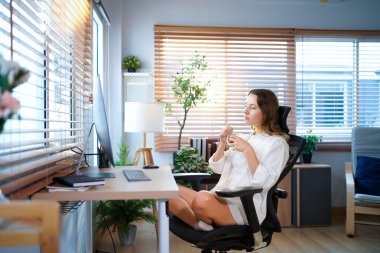 Beyaz bir kadın ofis masasında oturmuş masaüstü bilgisayarıyla kahve içiyor. Gülümseyen rahat genç bayan melez modern evdeki bilgisayarda çalışıyor..