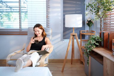 Genç kadın sıcak içecekten hoşlanıyor, kitap okuyor ve pencere ışığında rahat bir şekilde oturuyor..