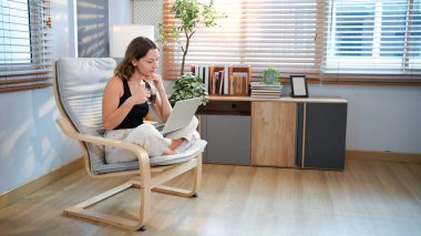 Kahve fincanı ve dizüstü bilgisayarı olan mutlu genç bir kadın evdeki oturma odasında oturuyor..