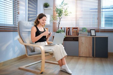 Kahve fincanı ve dizüstü bilgisayarı olan mutlu genç bir kadın evdeki oturma odasında oturuyor..