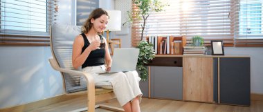 Kahve fincanı ve dizüstü bilgisayarı olan mutlu genç bir kadın evdeki oturma odasında oturuyor..