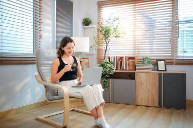 Kahve fincanı ve dizüstü bilgisayarı olan mutlu genç bir kadın evdeki oturma odasında oturuyor..