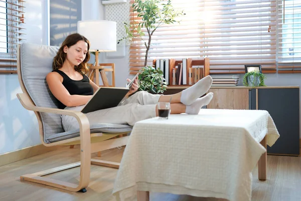 Genç kadın evde pencere kitap okuma oturma odasında rahatlatıcı önünde modern sandalye üzerinde oturan.