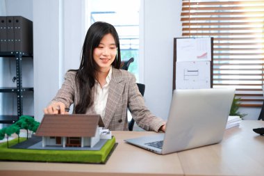 Gayrimenkul ile Ev Anlaşması, çekici, dengeli bir bayan ajan anlaşmanın ayrıntılarını araştırır, evrakları dikkatlice inceler. Yakınlarda bir model ev var, deneğin elle tutulur bir sembolü..