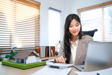 Ev modelli iş ipoteği emlakçısı, masada oturuyor, emlakçı hesap makinesi ve belgeler, ipotek ve mülk üzerinde çalışıyor..