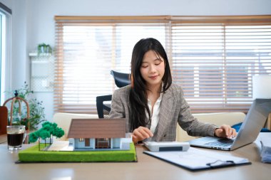 Ev modelli iş ipoteği emlakçısı, masada oturuyor, emlakçı hesap makinesi ve belgeler, ipotek ve mülk üzerinde çalışıyor..