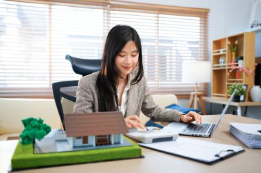 Gayrimenkul ile Ev Anlaşması, çekici, dengeli bir bayan ajan anlaşmanın ayrıntılarını araştırır, evrakları dikkatlice inceler. Yakınlarda bir model ev var, deneğin elle tutulur bir sembolü..