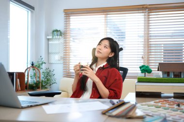 Genç mimar evde rahat bir atmosferde çalışıyor. Gülümseyen Asyalı kadın mimar masada sıcak kahve içiyor, ev ofisinin planlarıyla, bir fincan kahvenin tadını çıkarıyor..