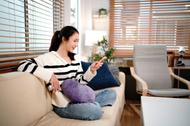 Güzel genç bir kadın televizyon seyrediyor. Rahat bir koltukta oturuyor. Elinde uzaktan kumandayla..