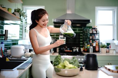 Beyaz spor kıyafetli bir kadın yeşil sebze ve meyve suyu hazırlıyor ve sağlıklı yaşam tarzında mutfakta kahvaltıda bir bardağa dolduruyor. Beslenme Konsepti.