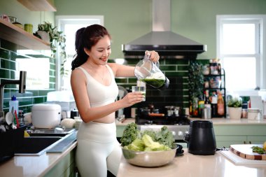 Beyaz spor kıyafetli bir kadın yeşil sebze ve meyve suyu hazırlıyor ve sağlıklı yaşam tarzında mutfakta kahvaltıda bir bardağa dolduruyor. Beslenme Konsepti.