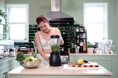Sağlıklı bir yaşam tarzı ve sağlık konsepti olan modern bir mutfakta sebzelerle karıştırılmış sağlıklı yeşil smoothie hazırlayan genç bir kadın..