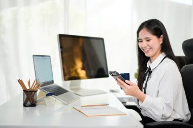 Mutlu Asyalı kadın akıllı telefon kullanıyor ve modern ofis masasında gülümsüyor. Cep telefonu tutan ve internetten e-posta ya da sosyal medyayı kontrol etmek ve aramak için sabah kahve içen Asyalı kadın.