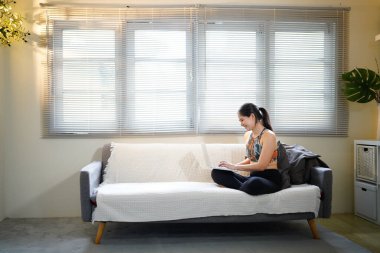 Spor giyimli bir kadın kanepede oturuyor, dizüstü bilgisayarla araştırma yapıyor ve internetten eğitim alıyor. O, spor ve egzersizlerde acemidir. Gülümsüyor ve mutlu hissediyor..