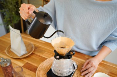 Genç Asyalı bir kadın, siyah çaydanlıktan kahve damlatıcısına sıcak su döküyor, modern mutfakta kadın kahvesi yapıyor, sabah yaşam tarzı, yavaş bar kafe ve ev yapımı konsept..