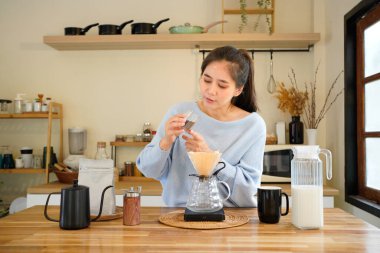 Genç Asyalı kadın barista evde taze sabah kahvesi yapıyor, kadın öğütücüden kahve dolduruyor, mutfakta kağıt filtreye döküyor, kahve yapıyor..