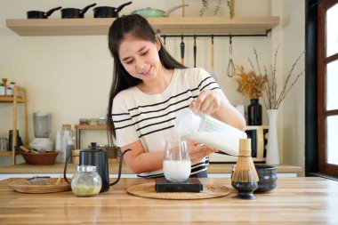 Evde modern mutfakta geleneksel Japon yeşil çayını hazırlayan ev yapımı bir matcha latte yapmak için bir sürahide taze süt döken bir kadın..
