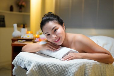 Bir sağlık merkezinde masaj masasında uzanmış kameraya bakan, siyah saçlı, güzel bir kadın..