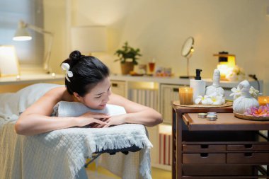 Güzel, esmer, sakallı bir kadın sağlık merkezinde masaj masasında yatarken mumlara ve masaj ekipmanlarına bakıyor..