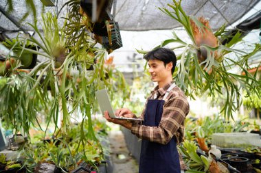 Asyalı bahçıvan, seranın içinde yağmur ormanları için egzotik bir tesiste çalışıyor. Kimlik tespiti için dizüstü bilgisayar kullanıyor, fabrika satışları planlıyor, envanteri kontrol ediyor, iş operasyonlarını yönetiyor..