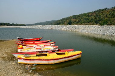 Colorful Kayaks Docked Along Pebble Shore by Calm River Amid Green Hills on a Sunny Day