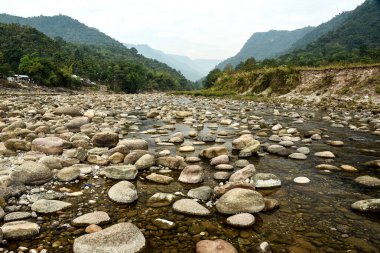 A calm river winds through a rocky bed, flanked by lush forests and majestic mountains. A small riverside settlement and a distant bridge add scale and charm to this natural landscape.