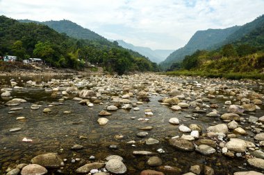 A calm river winds through a rocky bed, flanked by lush forests and majestic mountains. A small riverside settlement and a distant bridge add scale and charm to this natural landscape.